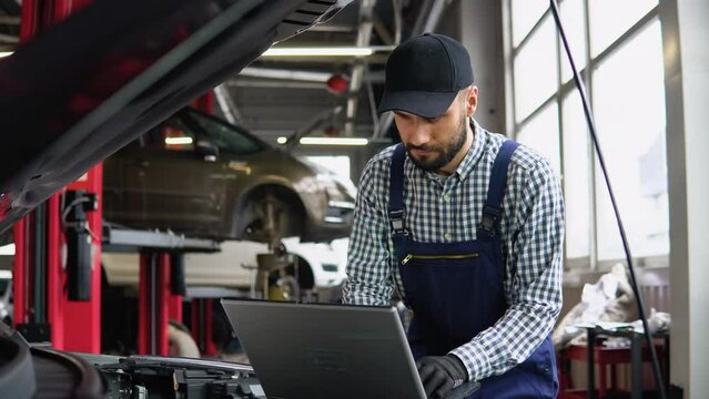 Experienced mechanic in uniform is using a laptop while repairing car in auto service