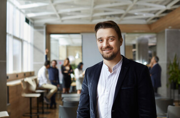 Portrait of smiling male executive director against background of office and workers. Successful Caucasian millennial man in shirt and jacket smiling looking at camera. Concept of business men.