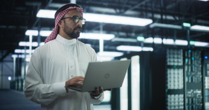 Saudi System Administrator Walks Between Rows of Operational Server Racks in Data Center. Engineer Uses Laptop Computer for Maintenance and Diagnostics in Cyber Security and Data Protection Facility