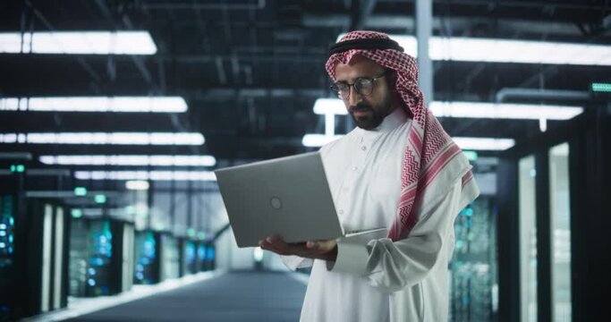 Portrait of a Middle Eastern IT System Engineer Wearing Traditional White Robe, Using Laptop Computer in a Modern Technological Server Farm Facility. Arab Artificial Intelligence Expert at Work