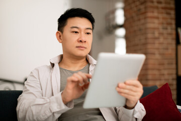 Middle aged korean man using digital tablet, surfing internet or shopping online at home, enjoying weekend pastime