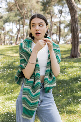 Fototapeta premium surprised woman with knitted sweater on shoulders holding daisy in summer park.