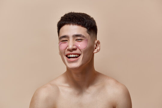 Portrait of handsome, smiling, young guy taking care after skin with under eye patches against light brown studio background. Concept of male beauty, skincare, cosmetology, men's health