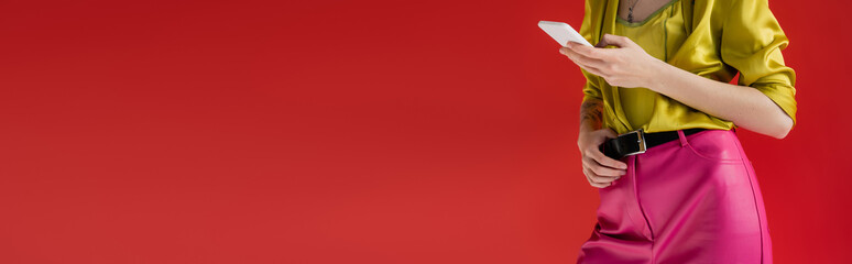 cropped view of tattooed woman in trendy outfit using smartphone on carmine pink background, banner.