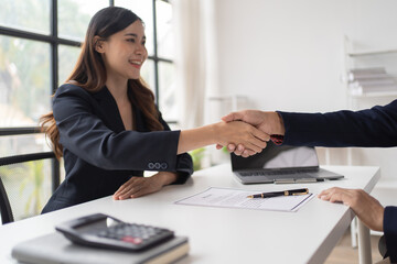 Happy young Asian businessman and woman shaking hands to congratulate successful collaboration on business deal with documents and laptop on table in the office.