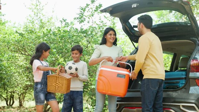 Happy Indian couple with kids taking out bags and picnic bucket from car boot - concept of summer holidays, togetherness and family vacation
