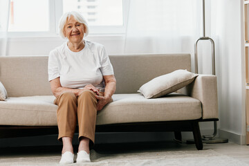 Obraz na płótnie Canvas elderly woman sits on a sofa at home against the backdrop of a window and a happiness smile, stylish interior. Lifestyle retirement.