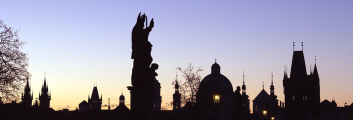 Sihouette of saint statue and skyline of Prague's old town with church towers on the famous Charles Bridge at dawn. 