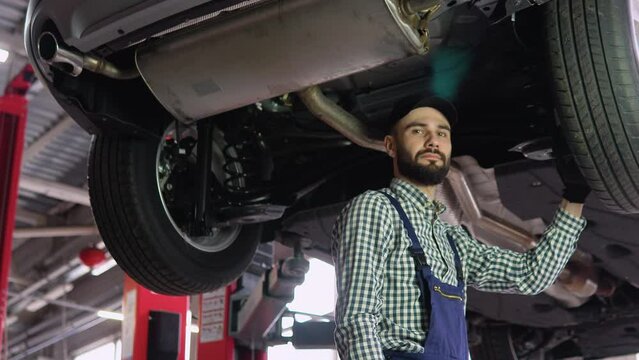 Professional Repairman is Wearing Gloves and Using a Ratchet Underneath the Car. Portrait Shot of a Handsome Mechanic Working on a Vehicle in a Car Service. Modern Clean Workshop