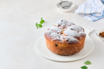 Twisted yeast cake with raisins sprinkled with powdered sugar on a white ceramic board against a light concrete background. Easter concept.