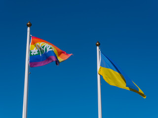 The flag of Ukraine and the rainbow flag on the flagpoles on the background of a sky. Two flags and a cloudless sky.