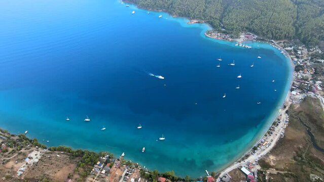 Aerial drone video showcasing the picturesque Akb&uuml;k Cove in Akyaka, Muğla, Turkey, featuring yachts and boats anchored in the pristine waters surrounded by a scenic coastline, mountains, and trees.