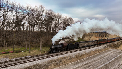 Obraz premium A Drone View of a Steam Locomotive Approaching Traveling Thru Fields and Meadows, Blowing White Smoke on a Winter Day
