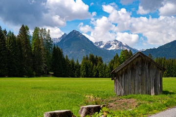 View of mountain peaks over lush green meadows and forests with shed in foreground on sunny day with blue sky and white clouds