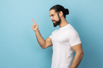 Side view of angry man with beard wearing T-shirt pointing finger at camera and looking with dissatisfied suspicious expression, warning about troubles. Indoor studio shot isolated on blue background.