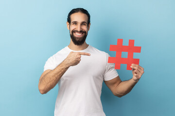 Portrait of happy man with beard wearing white T-shirt holding and pointing hashtag symbol,...