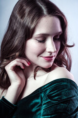 Naklejka premium Beauty portrait of pleased smiling beautiful woman with closed eyes wearing green dress, keeps hand near her face, having gently look. Indoor studio shot isolated on gray background.