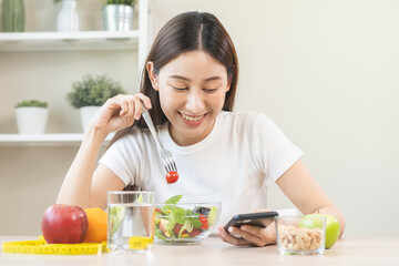 Obraz na płótnie Canvas Dieting, smile asian young woman eating, holding fork at tomato, diet plan nutrition with fresh vegetables salad, enjoy meal while using smartphone. Nutritionist of healthy, nutrition of weight loss.