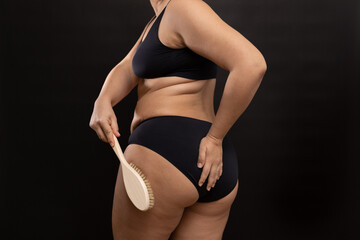 Fat woman in black underwear massaging hips with brush, back view. Flaunt figure imperfections. Dry anti cellulite massage. Studio portrait over black background. Concept of skin care, body positive.