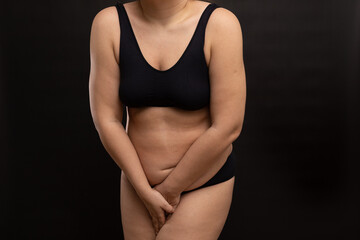 Plus size woman in black underwear covering with hands naked body. Need to go to toilet. Studio portrait over black background. Concept of human biological needs, body positive, patience.