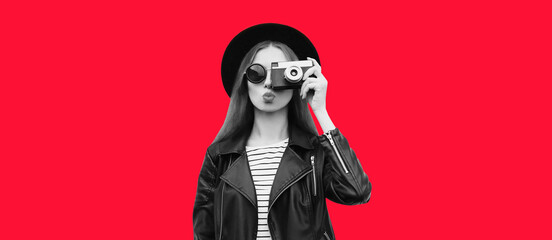 Portrait of beautiful stylish young woman photographer with film camera wearing black round hat, leather jacket isolated on red background