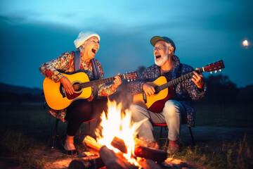 Old couple of husband and wife, enjoying retirement - they are singing and playing guitar by a campfire at blue hour while on a trip. Generative AI - obrazy, fototapety, plakaty
