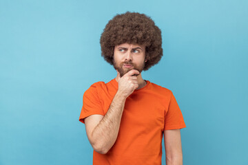 Portrait of man with Afro hairstyle wearing orange T-shirt holding chin with hand, need to think,...