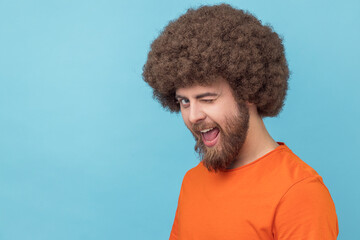 Portrait of flirting man with Afro hairstyle wearing orange T-shirt in good mood, smiling broadly and winking at camera with toothy smile. Indoor studio shot isolated on blue background. - obrazy, fototapety, plakaty