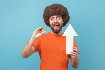 Portrait of happy positive excited man with Afro hairstyle wearing orange T-shirt showing white arrow pointing up and bitcoin, growth. Indoor studio shot isolated on blue background.