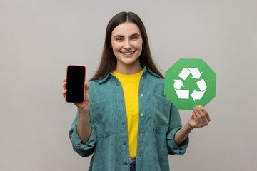 Smiling positive woman showing mobile phone with blank screen and recycling symbol, being happy,...