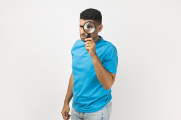 Portrait of unshaven man in blue T- shirt standing standing, holding magnifying glass and looking...