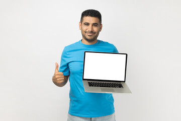 Portrait of delighted smiling attractive unshaven man wearing blue T- shirt standing showing blank screen of laptop, showing like gesture, thumb up. Indoor studio shot isolated on gray background.