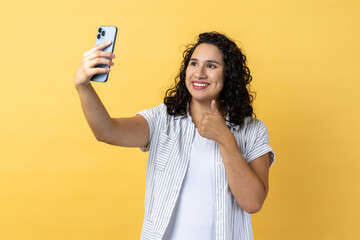 Portrait of satisfied beautiful woman with dark wavy hair holding mobile phone making selfie or...
