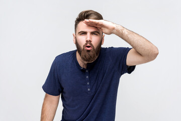 Portrait of amazed man with beard wearing dark blue T-shirt looking far away at distance with hand...