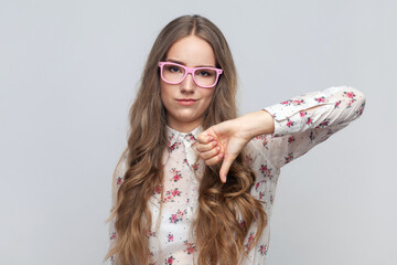 Portrait of disappointed young adult woman in glasses with long blond hair showing thumb down,...