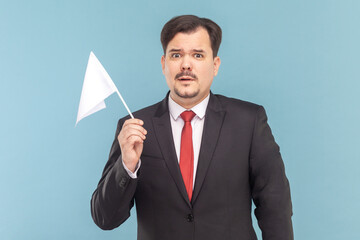 Portrait of man with mustache standing holding a white flag, looking at camera with scared expression, wearing black suit with red tie. Indoor studio shot isolated on light blue background.