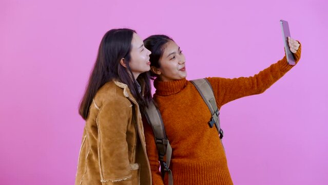 4K, Two Asian female tourists wearing long sleeves, Travel in different places, bring tablet to take pictures of two people, look at pictures taken together, Isolated indoor studio on pink background.