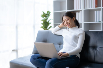 Happy young Asian woman relaxing using laptop computer on sofa at home.