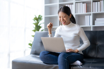 Happy young Asian business woman successful excited raised hand rejoicing with laptop at home.