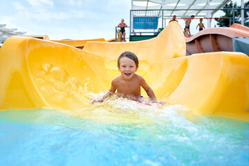 Cheerful blue-eyed kid on a water slide in the water park, a little boy merrily slides into the...