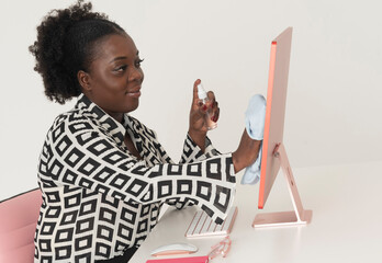 England, UK. 2023. African office worker spraying a computer screen and cleaning with a cloth....