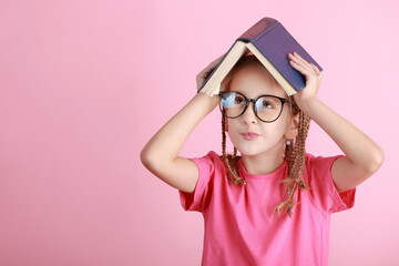 Back to school concept. Smiling pretty little school girl in glasses is holding book over head like roof isolated on pink background. Childhood lifestyle concept. Education in school. September 1