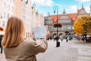 Back view of attractive young female tourist holding a paper map on central square in Gdansk old town. Traveling Europe in autumn. High quality photo