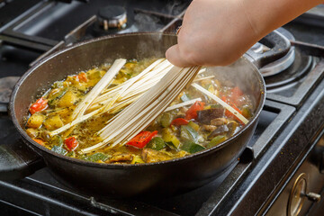 A woman's hand puts udon in a pan with stewed vegetables.