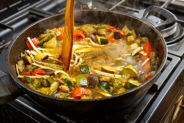 A wooden spatula mixes udon with stewed vegetables in spices in a frying pan.