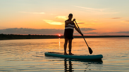 A man in shorts on a SUP board with a paddle at sunset swims in the water of the lake in the glare from the setting sun.