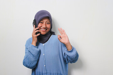 Happy young Asian girl in hijab dressed in casual shirt say hello to the camera while talking on phone