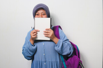 Young Asian girl student in hijab covering her face with book isolated on white background.