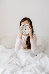 8 years girl holding alarm clock 7 am hidding the face, sitting on bed under white blanket, sleep at morning. Time go to school, wake up early at home