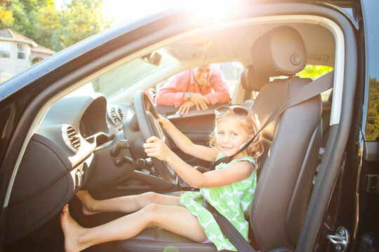 Little girl sitting in driver's seat of car wearing seatbelt, pretending to be old enough to drive and showing she knows the importance of a seat belt as her smiling father watches on on a sunny summe